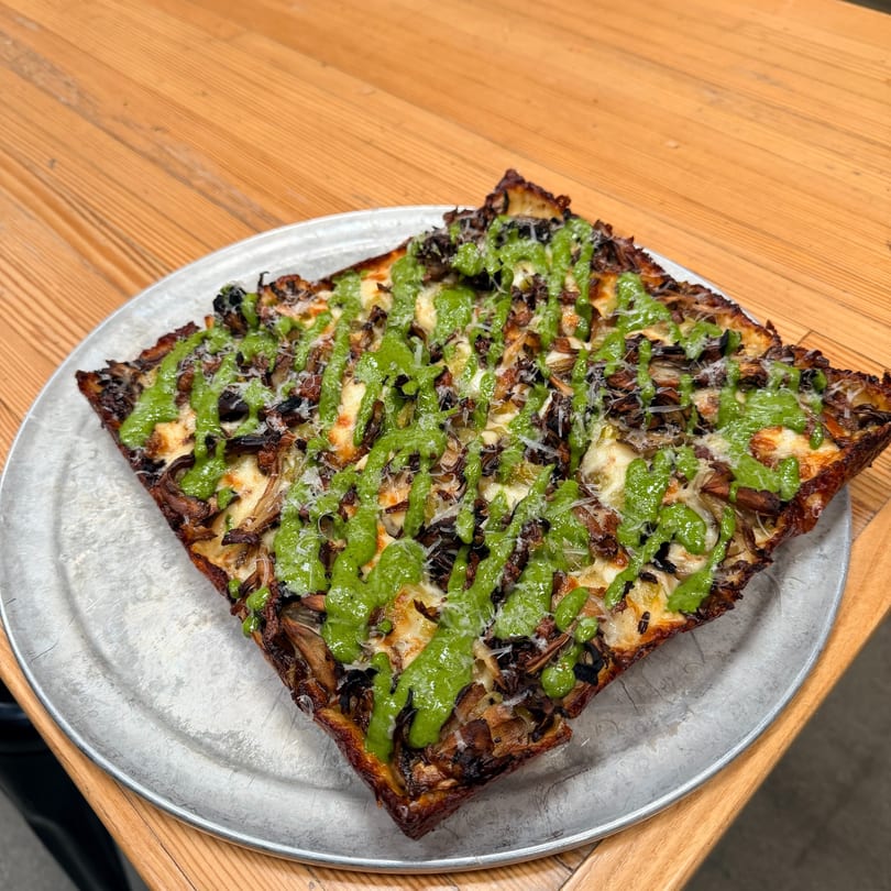 Square Detroit-style pizza with mushrooms, cheese, and a vibrant green pesto drizzle on a metal tray.