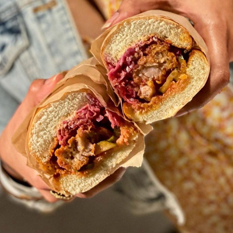 Hands holding a large sandwich, cut in half, revealing fried chicken, red cabbage, and sauce.