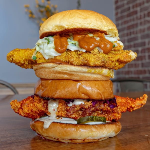 Two stacked fried chicken sandwiches with coleslaw, sauce, and pickles on a wooden table.