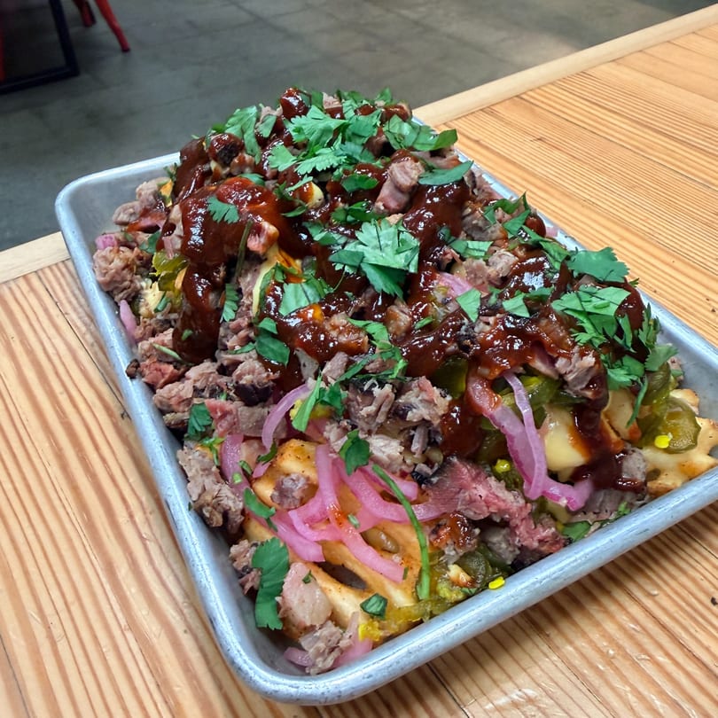 A metal tray of waffle fries topped with pulled pork, BBQ sauce, pickled onions, and cilantro.